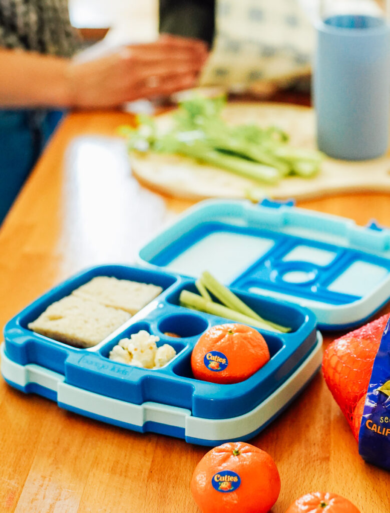 School lunch in a blue lunchbox with a sandwich, orange, celery sticks, and popcorn.