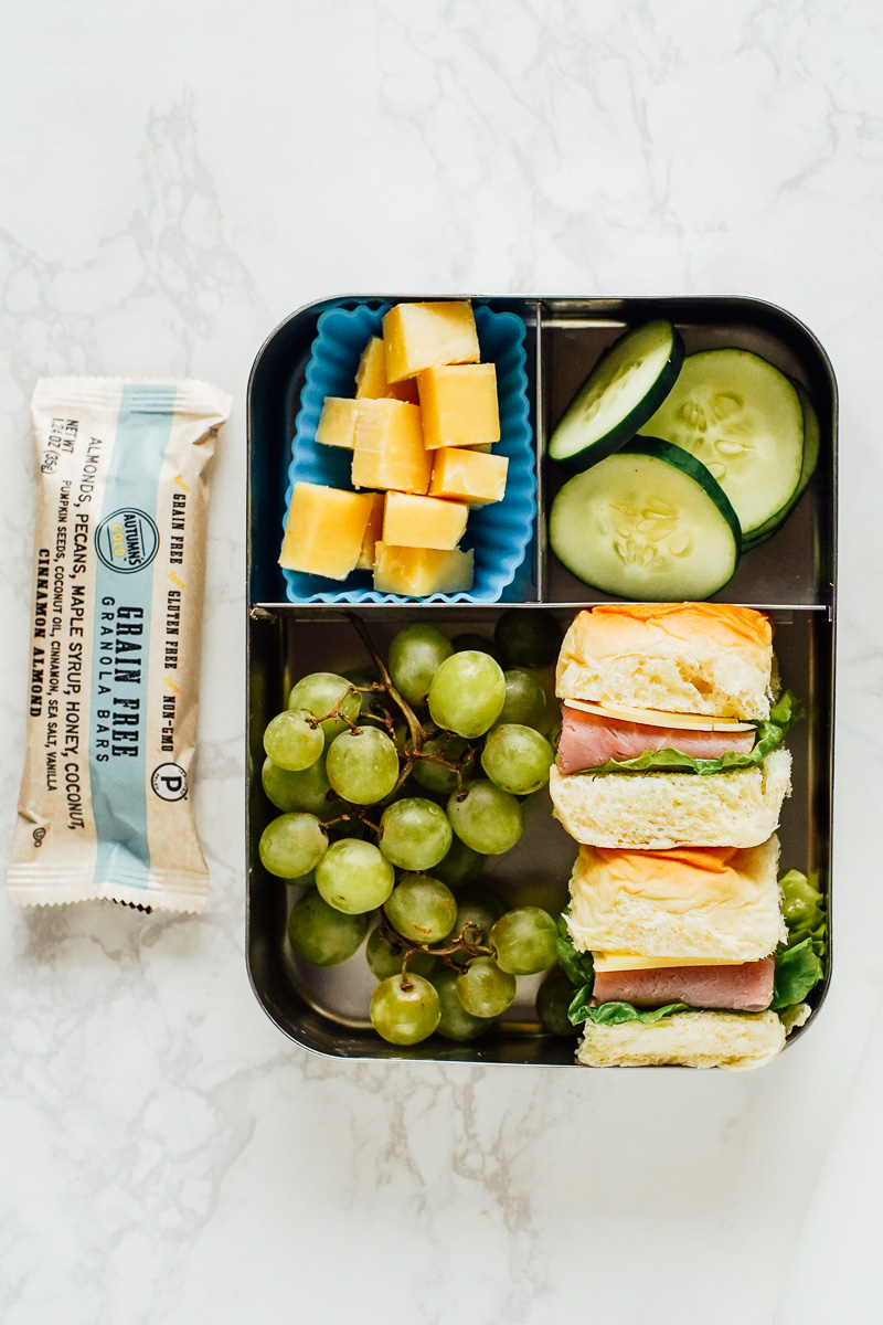 Two sandwiches with turkey or ham and cheese, grapes, cheese cubes, cucumber slices, and granola bar in a bento box lunchbox.