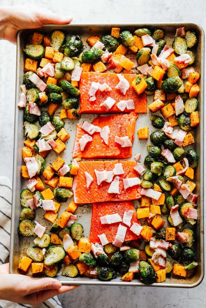 Roasted veggies pushed to the side of the sheet pan with raw salmon in the center topped with bacon.
