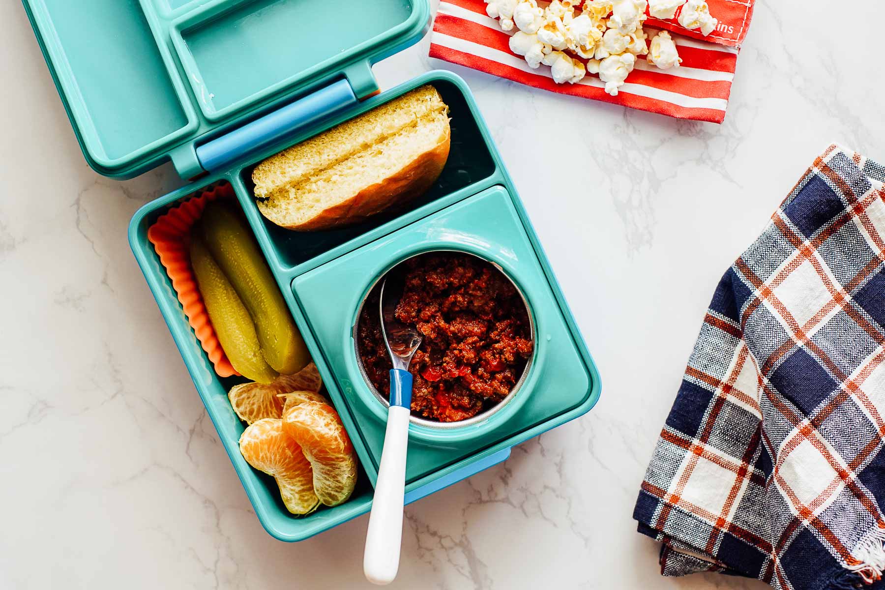 Leftover sloppy joe meat in a thermos with a bun, orange slices, pickle, and popcorn on the side in a lunchbox.