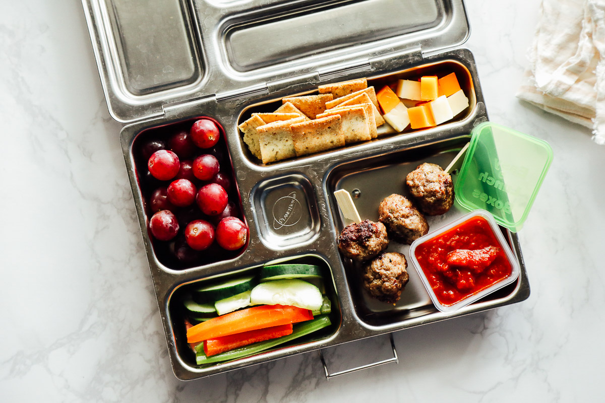 Meatballs on a skewer with marinara sauce, carrots and cucumbers, cheese and crackers, and grapes in a lunchbox.