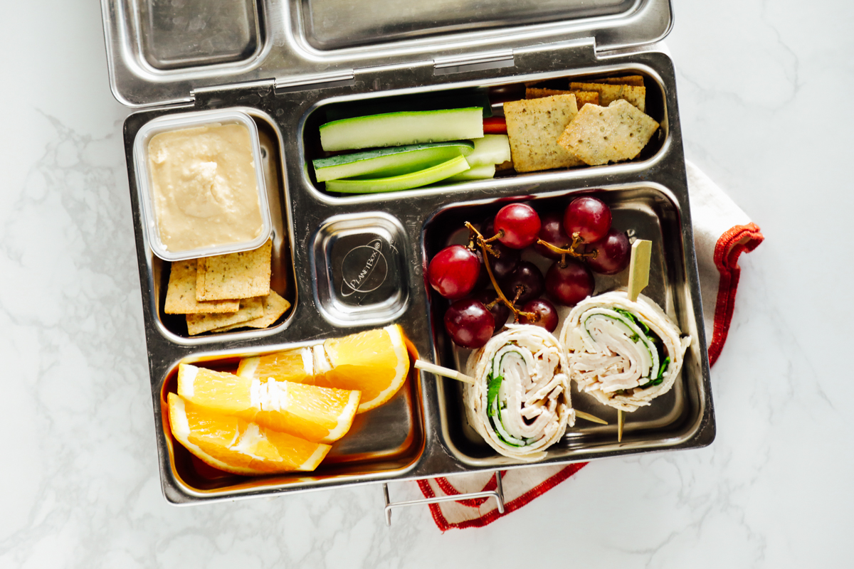 Turkey pinwheels with oranges, hummus and crackers, and celery sticks in a metal lunchbox.